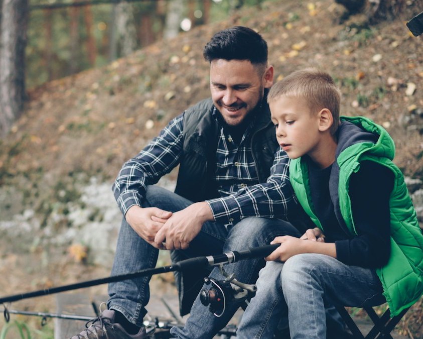 A father and his son sitting together by a lake while fishing, smiling and talking