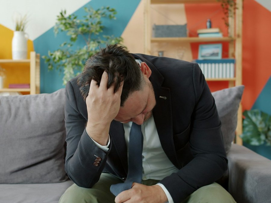 A middle-aged man in a blazer sitting on a couch with his head in his hand, looking stressed