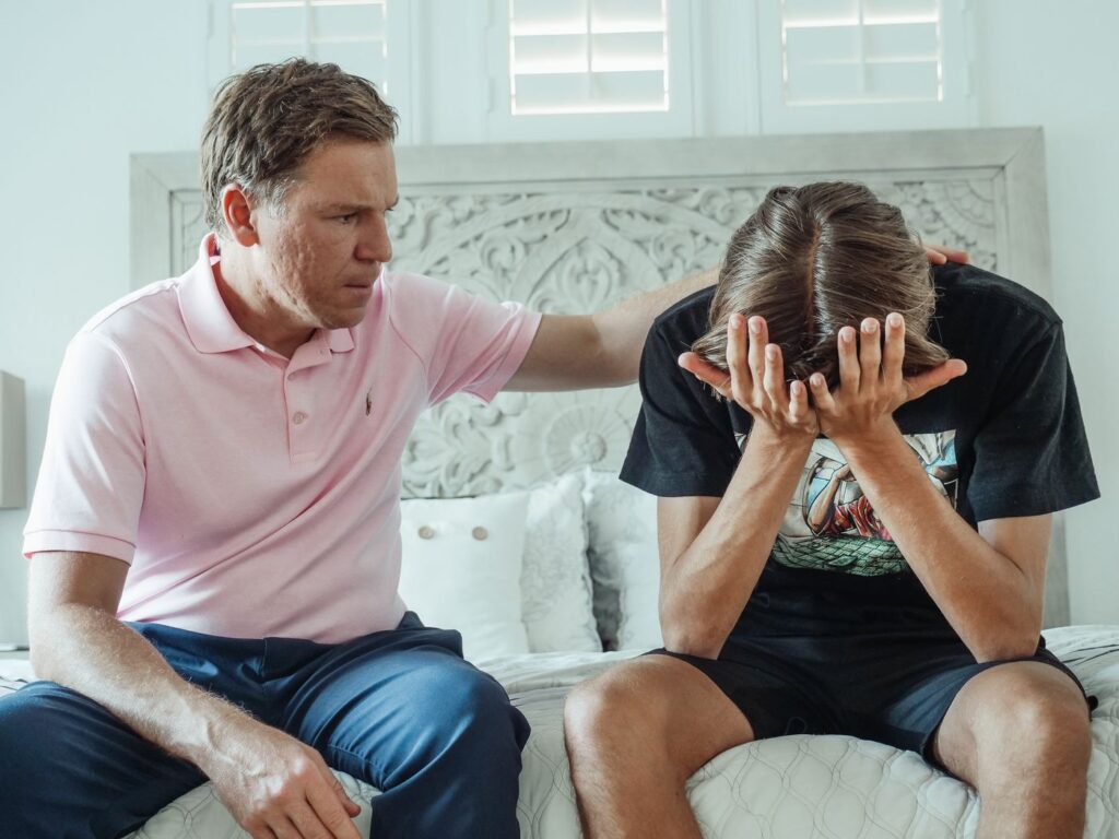 Father comforting his teenage son, who sits with his head in his hands